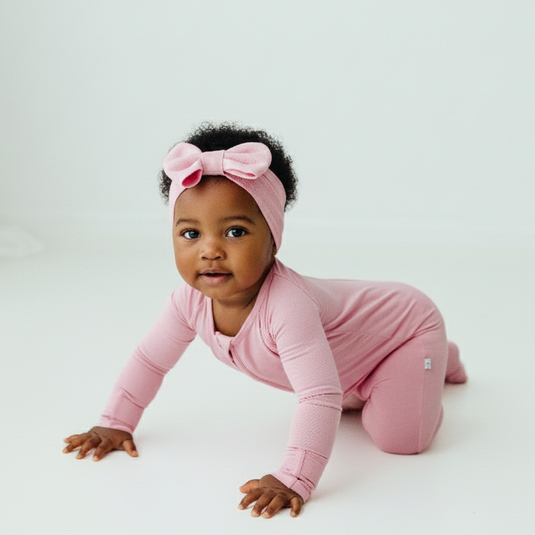 baby girl in Petal Glow bamboo baby sleeper – highlighting the luxurious stretch and gentle touch. These baby sleepers are a must.-have for every new parent with a baby girl. Crawling with pink bow in hair.