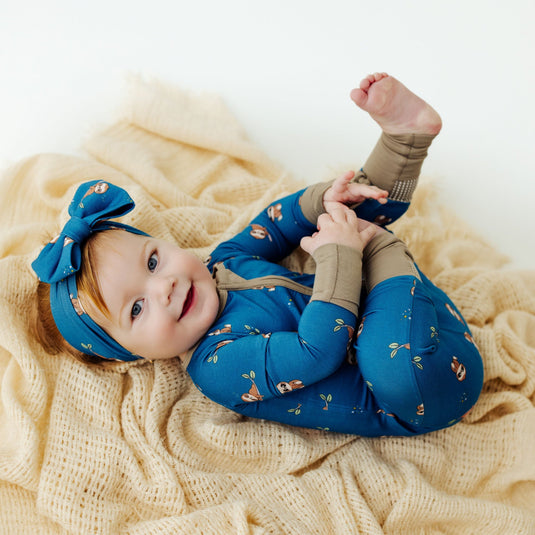 Baby lying on a beige blanket wearing a blue bamboo sleeper and headband.