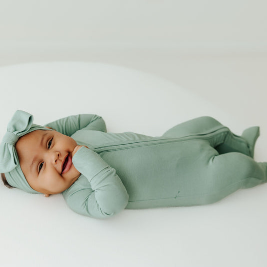 Baby laying down in a Frosted Sage bamboo sleeper and one of our matching baby bows. A gender-neutral favorite in bamboo baby clothes and perfect for baby shower gifts.